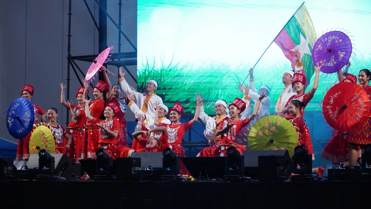 A Myanmar performance group in red costumes raising parasols and a flag on stage